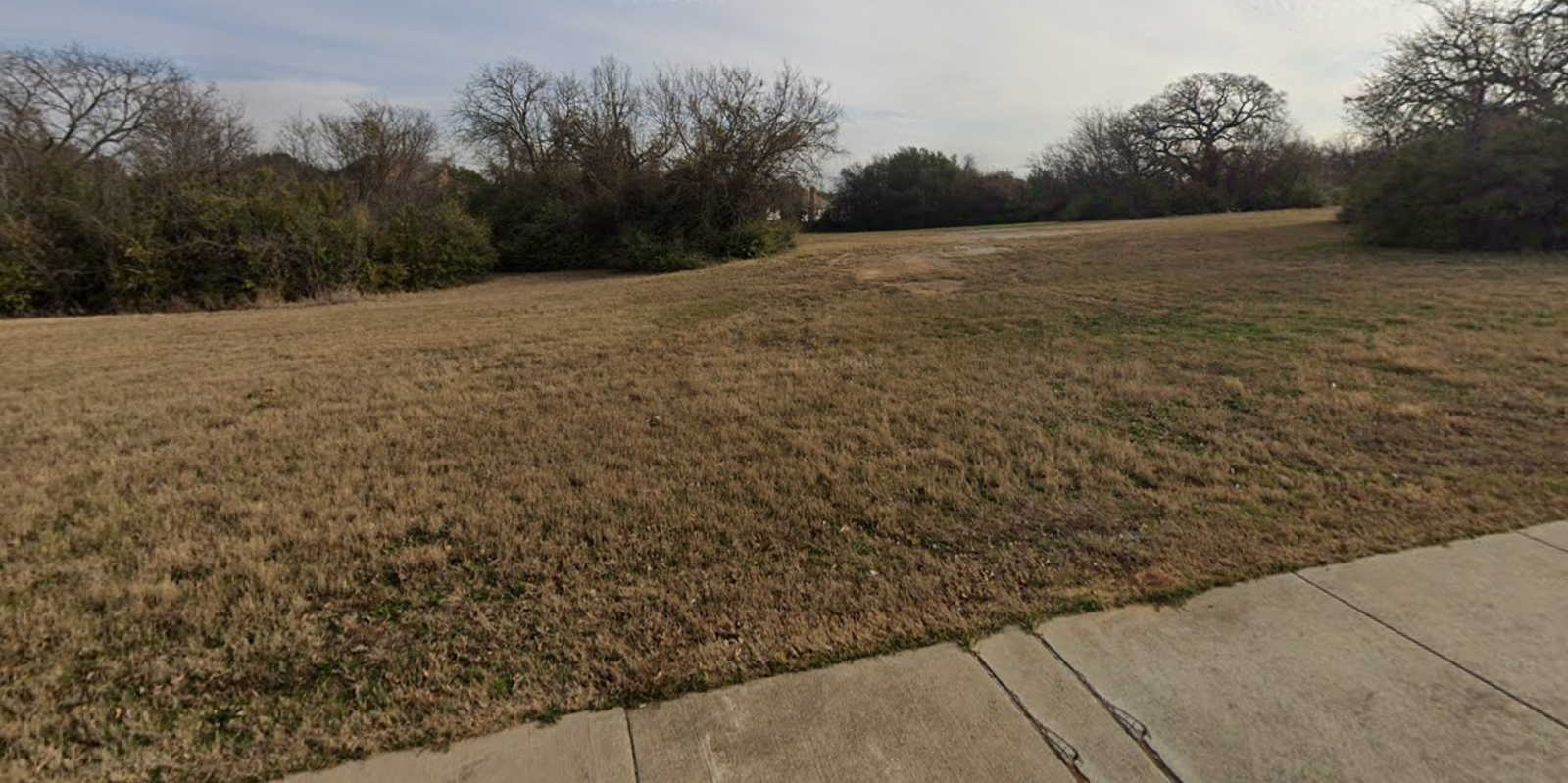 Grassy Norman Lee Robinson Park site in Arlington, bordered by trees and a sidewalk