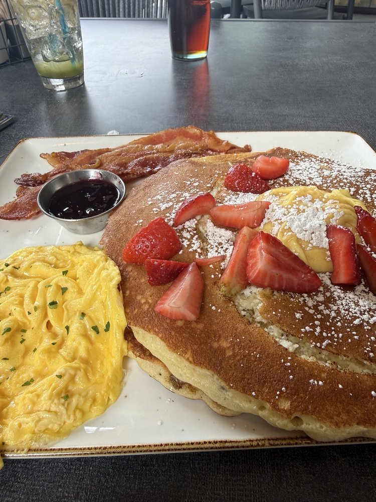 First Watch Pancake Breakfast First Watch breakfast plate with strawberry-topped pancakes, scrambled eggs, and bacon