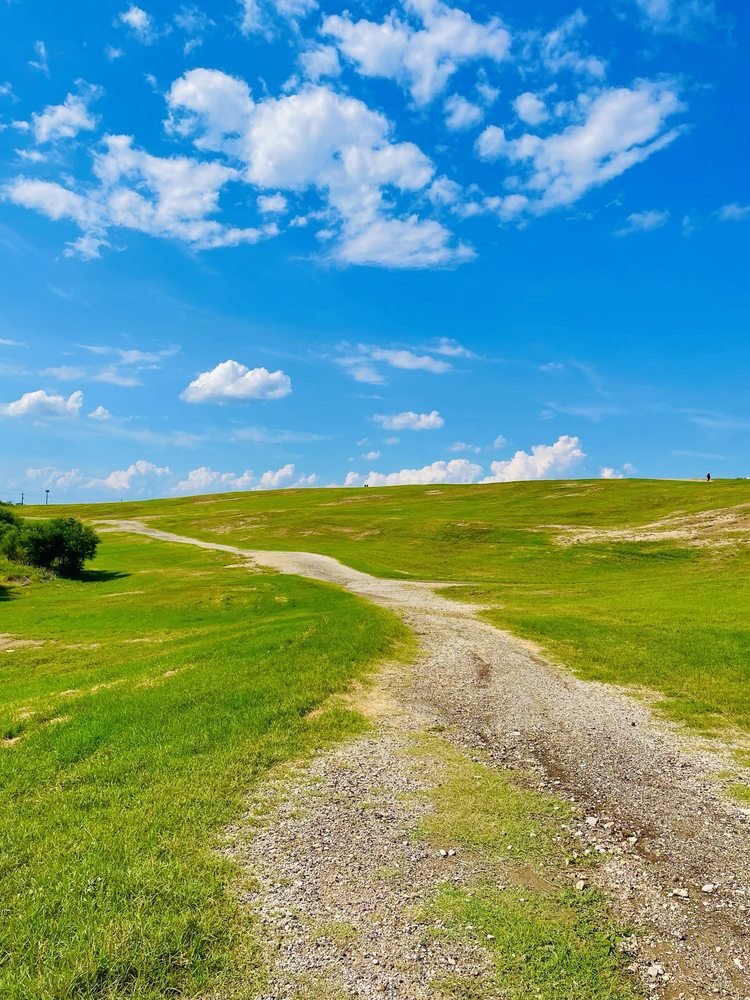 Gravel trails cross grassy hills at Pearsall Park beneath a blue sky.