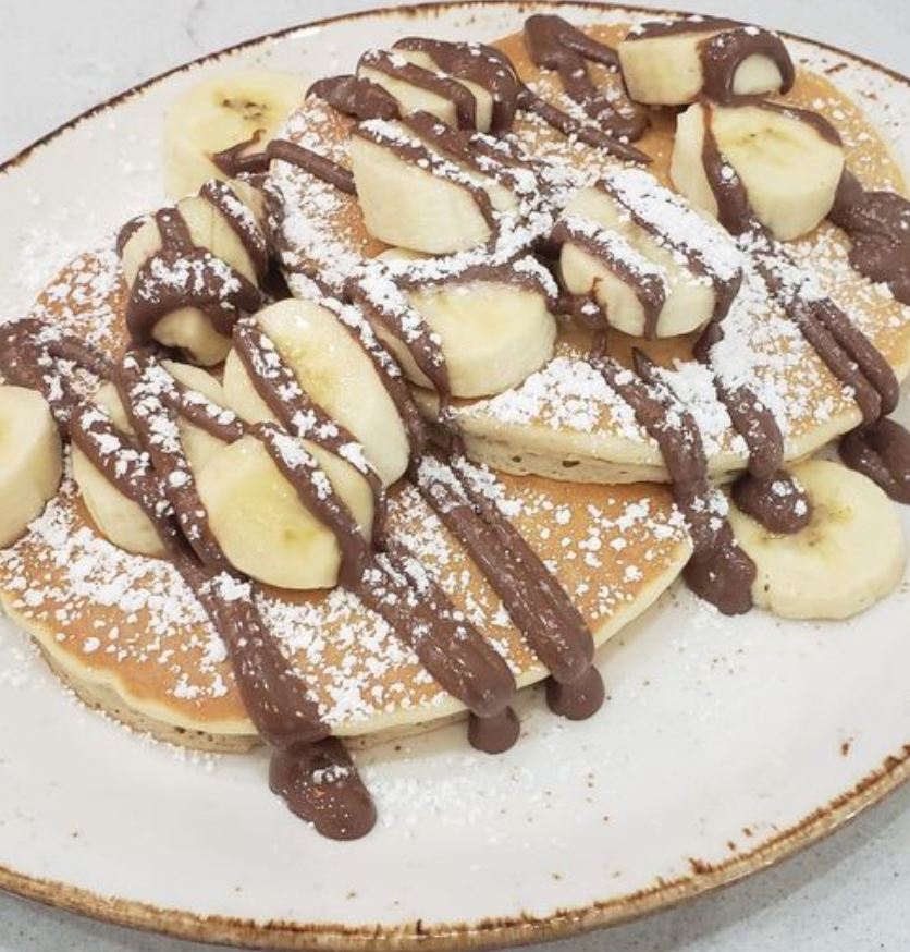 Pancakes topped with bananas, chocolate-colored drizzle and powdered sugar, representative food for the Brunch District Celina story