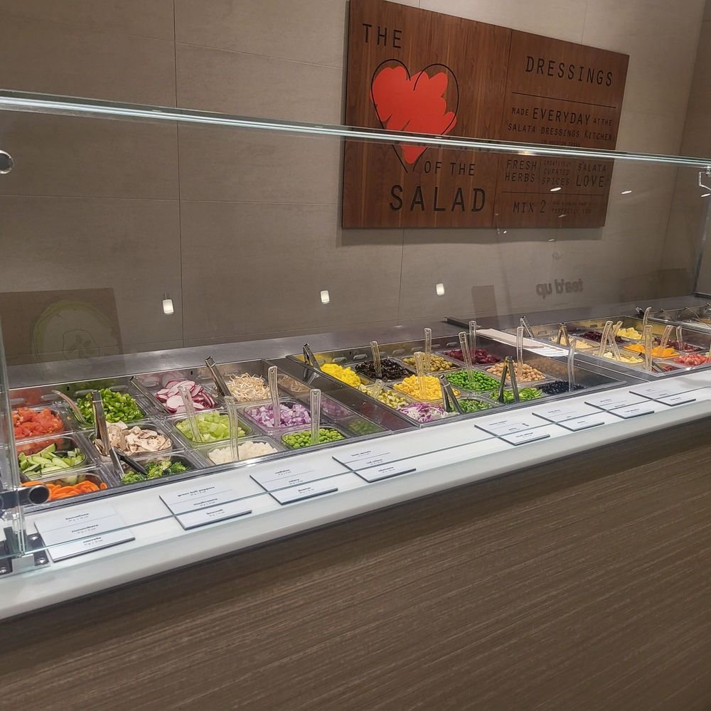 Salata salad assembly counter Fresh vegetables and toppings along a Salata salad assembly counter