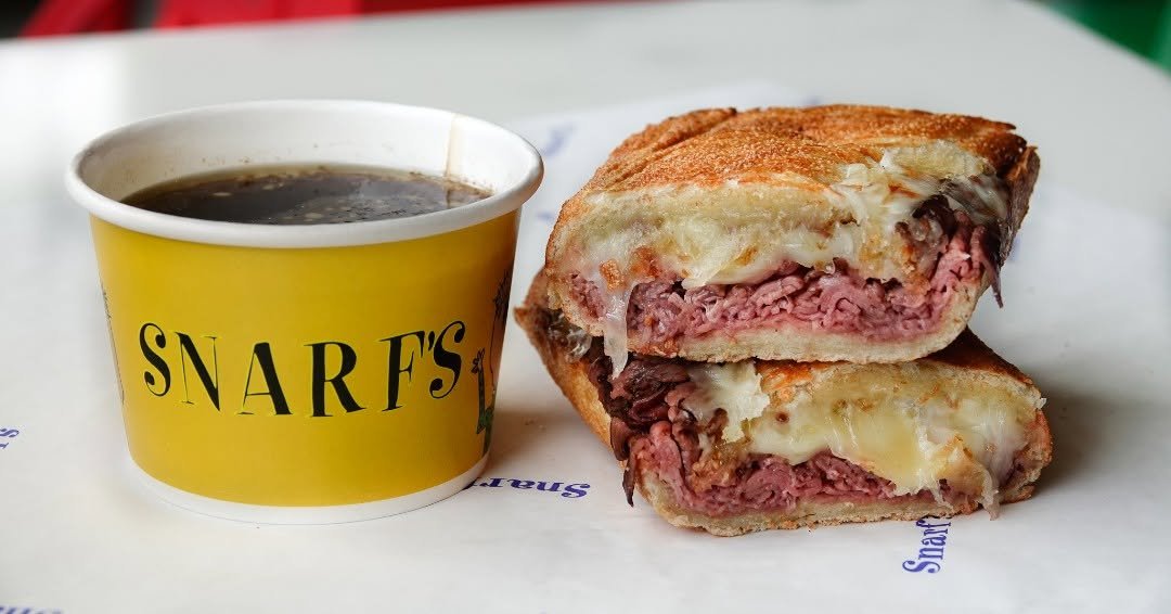 Snarf's toasted sandwich with sliced meat and melted cheese beside a cup of dipping broth