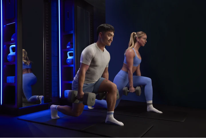 Two people performing lunges with dumbbells in a blue-lit fitness studio